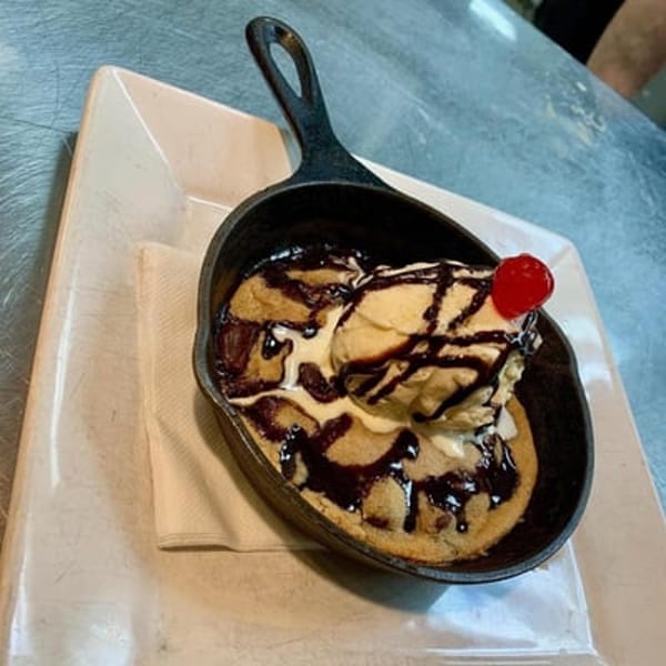 A warm chocolate chip cookie skillet topped with vanilla ice cream, chocolate syrup, and a cherry.